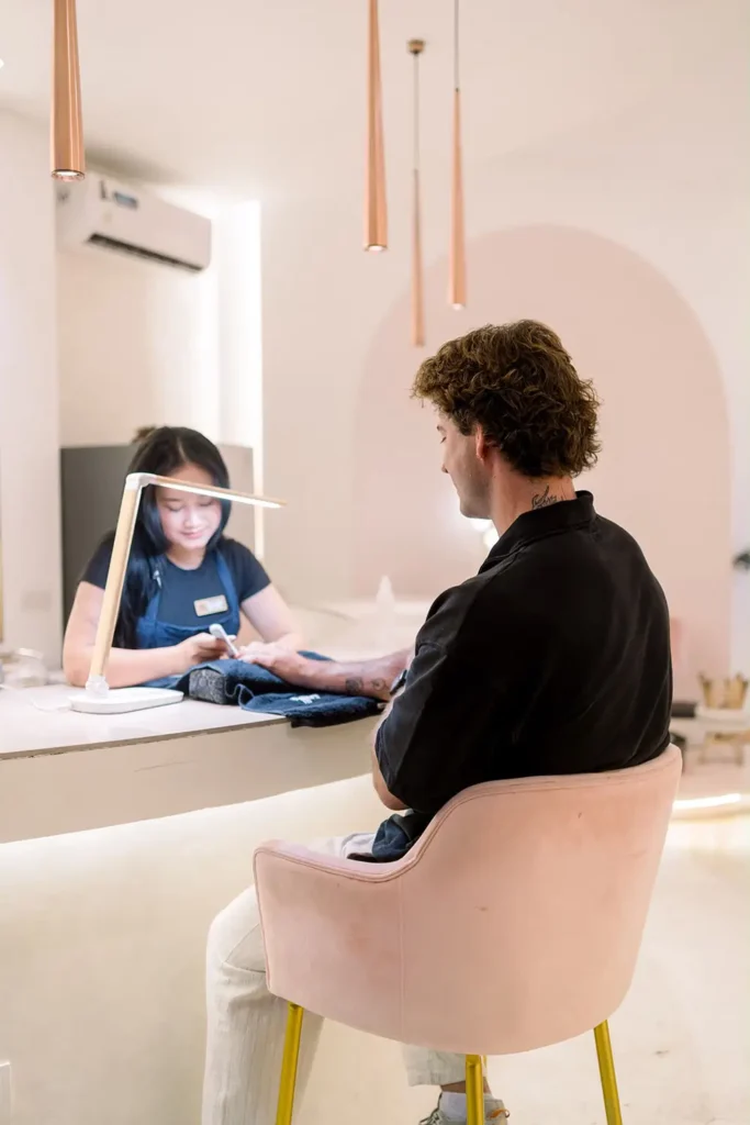 A male client receiving a manicure from a female technician at a brightly lit white counter, highlighting the services of a nail salon Bali for ladies and gentlemen located at Moiselle one stop beauty Canggu salon.